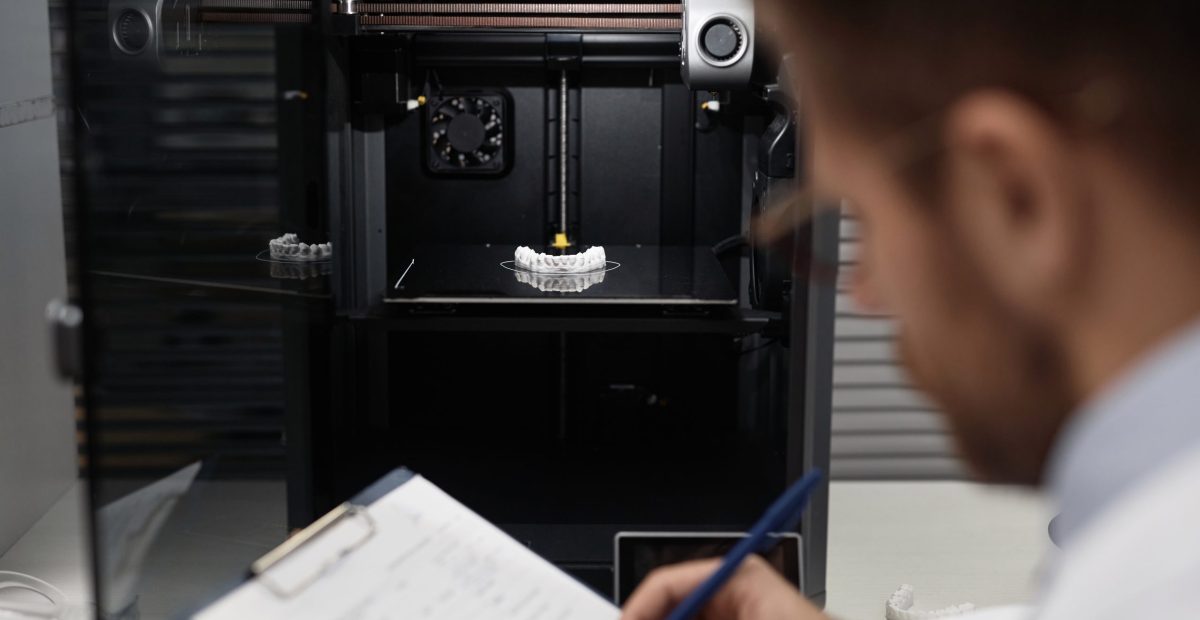 Technician Documenting Printed 3D Dental Prosthesis Medium shot of illuminated toothed jaw bone model on hot bed of enclosed 3D printer, unrecognizable male technician filling in paper in blurred foreground